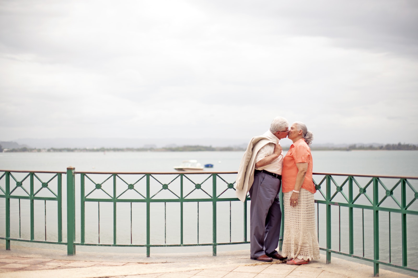 Side view full body romantic senior couple wearing trendy outfits standing on waterfront promenade and kissing gently on overcast weather