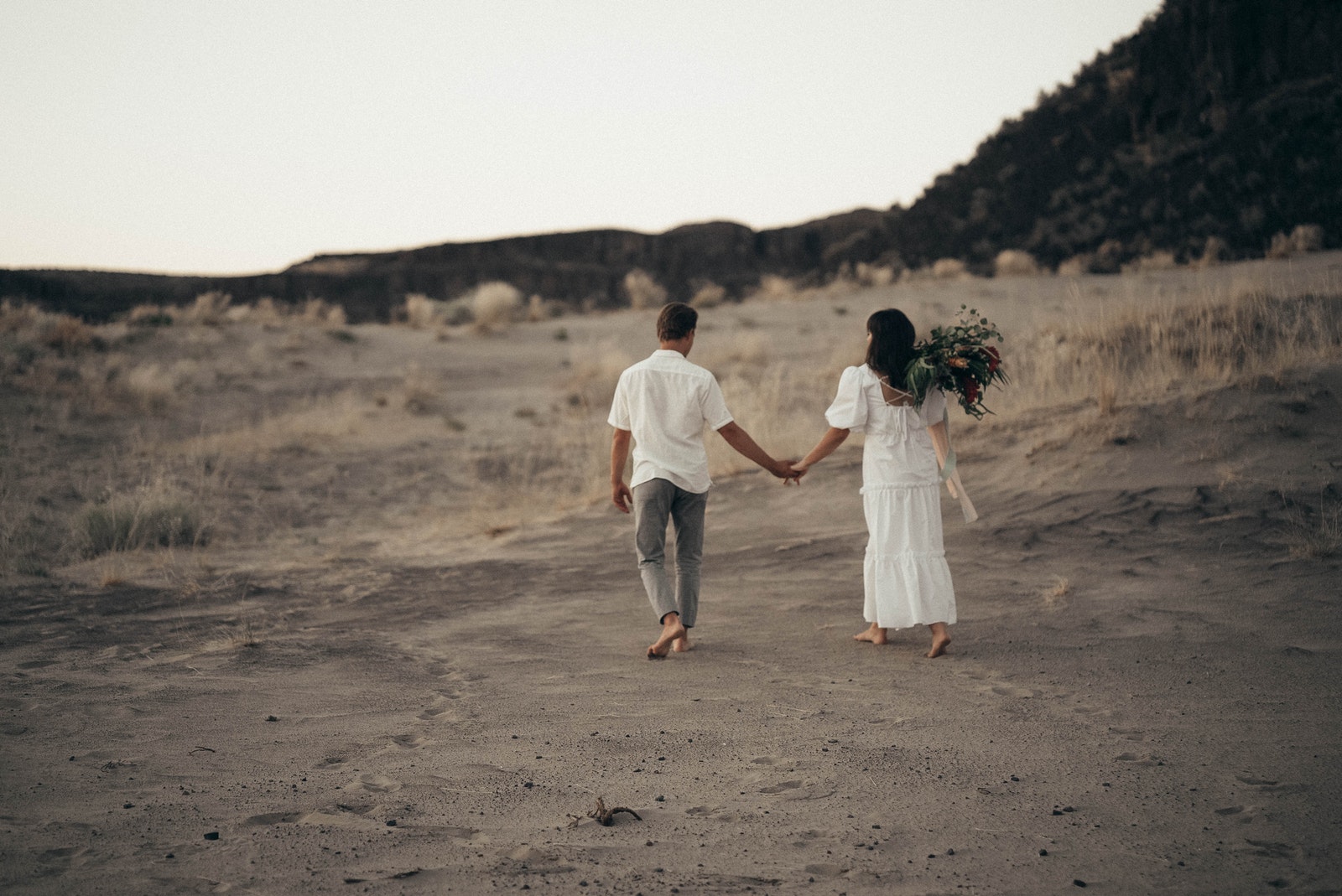 Full body back view of anonymous bride with flowers and groom holding hands while strolling on sandy field during wedding celebration