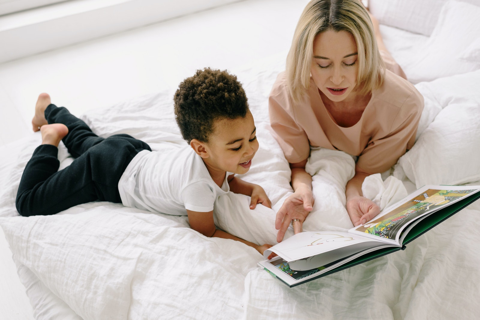 Mother and Son Reading Book Together while Lying on a Bed with White Blanket